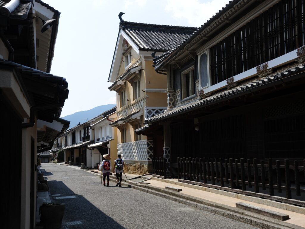Two travelers in woven sedge hats stroll past ochre plaster merchant houses in Uchiko, where careful restoration keeps the Edo and Meiji streetscape alive.