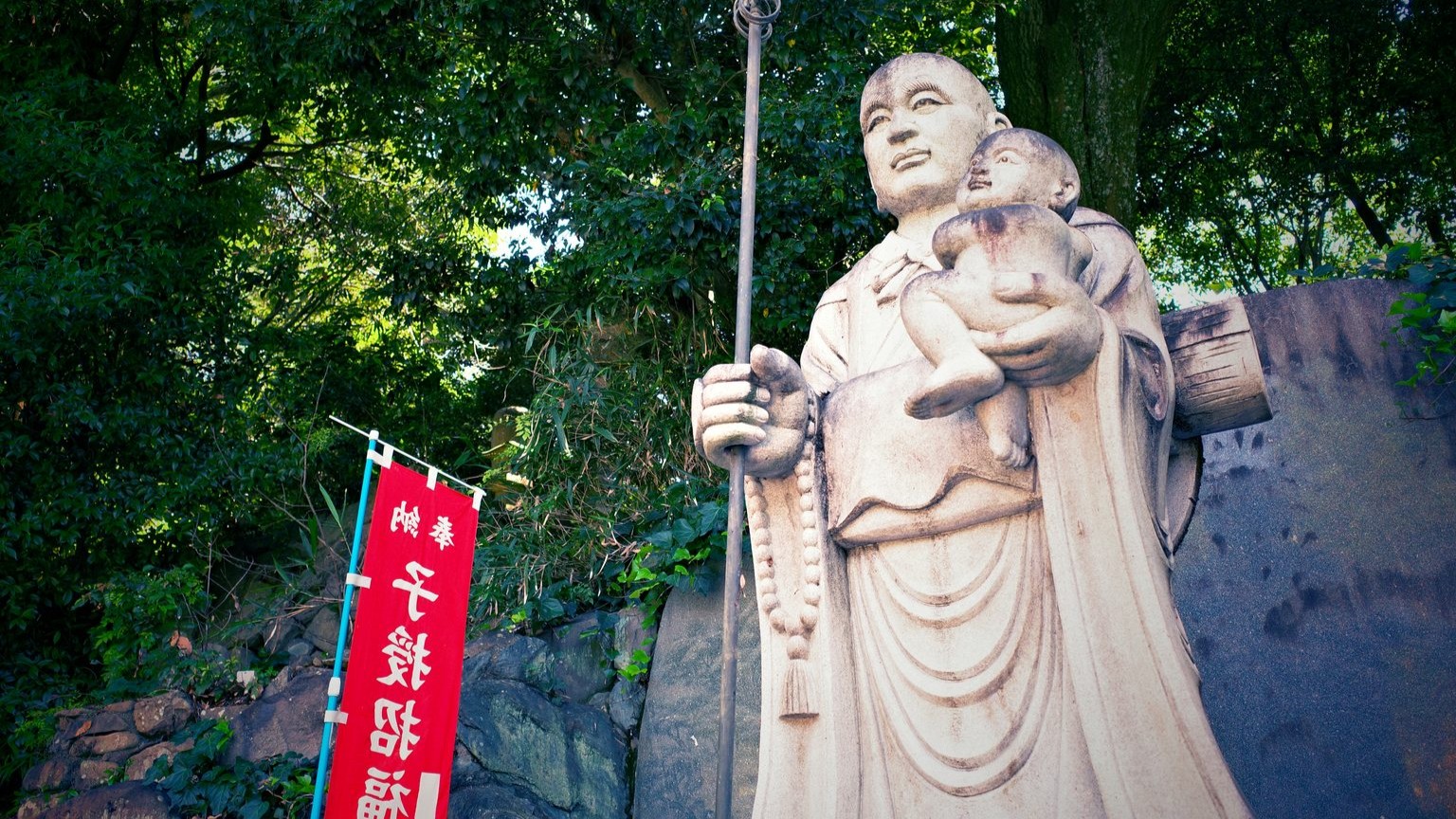 A stone statue of Kūkai holding a child along with a red flag that reads "the Great Teacher who bestows children and invites good fortune."