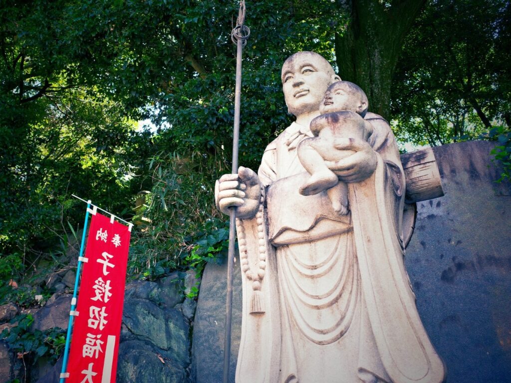 A stone statue of Kūkai holding a child along with a red flag that reads "the Great Teacher who bestows children and invites good fortune."