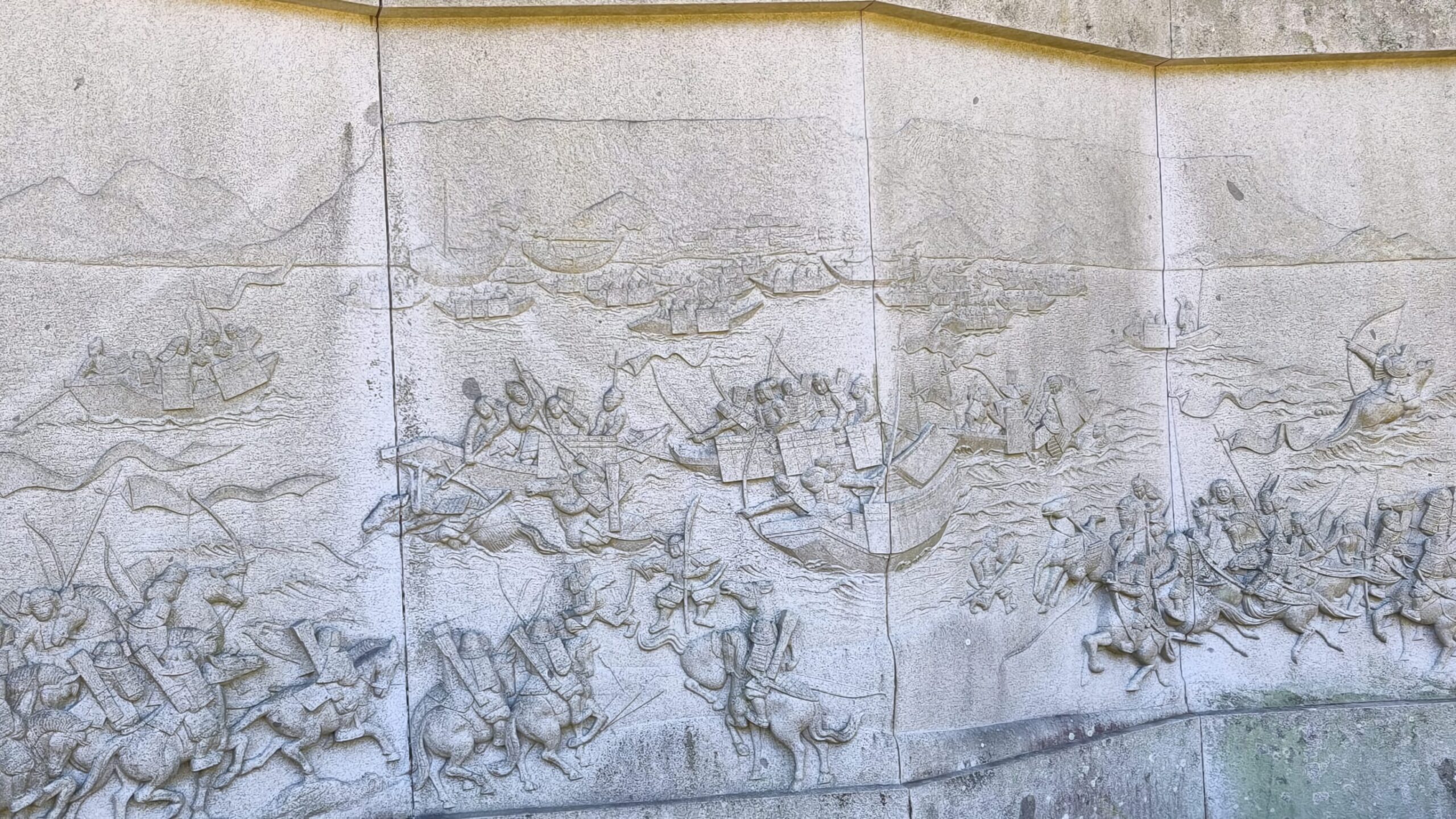 A stone relief depicting the battle of Yashima—horse-mountain samurai warriors on the shore watch the action play out between the legendary heroes of the battle in the bay beneath the mesa