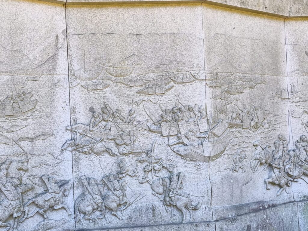 A stone relief depicting the battle of Yashima—horse-mountain samurai warriors on the shore watch the action play out between the legendary heroes of the battle in the bay beneath the mesa