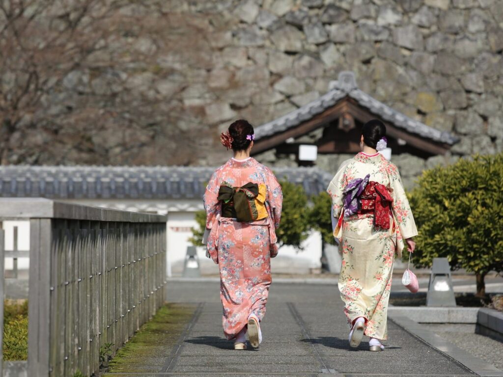 Two kimono-clad women walk through the gardens of the Ninomaru park at Matsuyama castle. Citrus trees and concrete squares mark the original position of demolished buildings. The stone wall of the castle looms in the background.